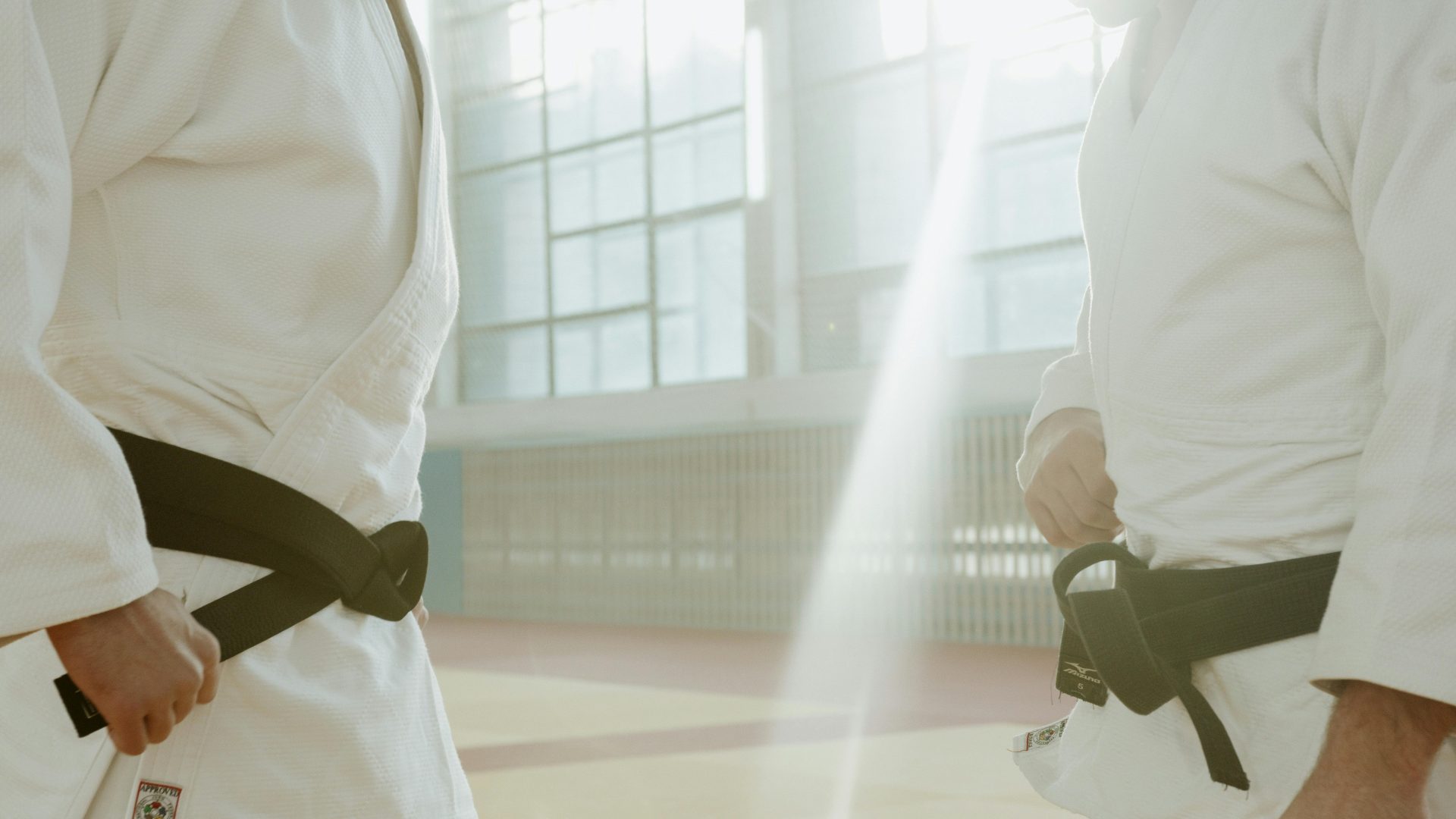 Dos artistas marciales con uniformes blancos se preparan para entrenar en un gimnasio cubierto bien iluminado.